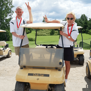 Sue lusk and other person standing on golf cart waving.